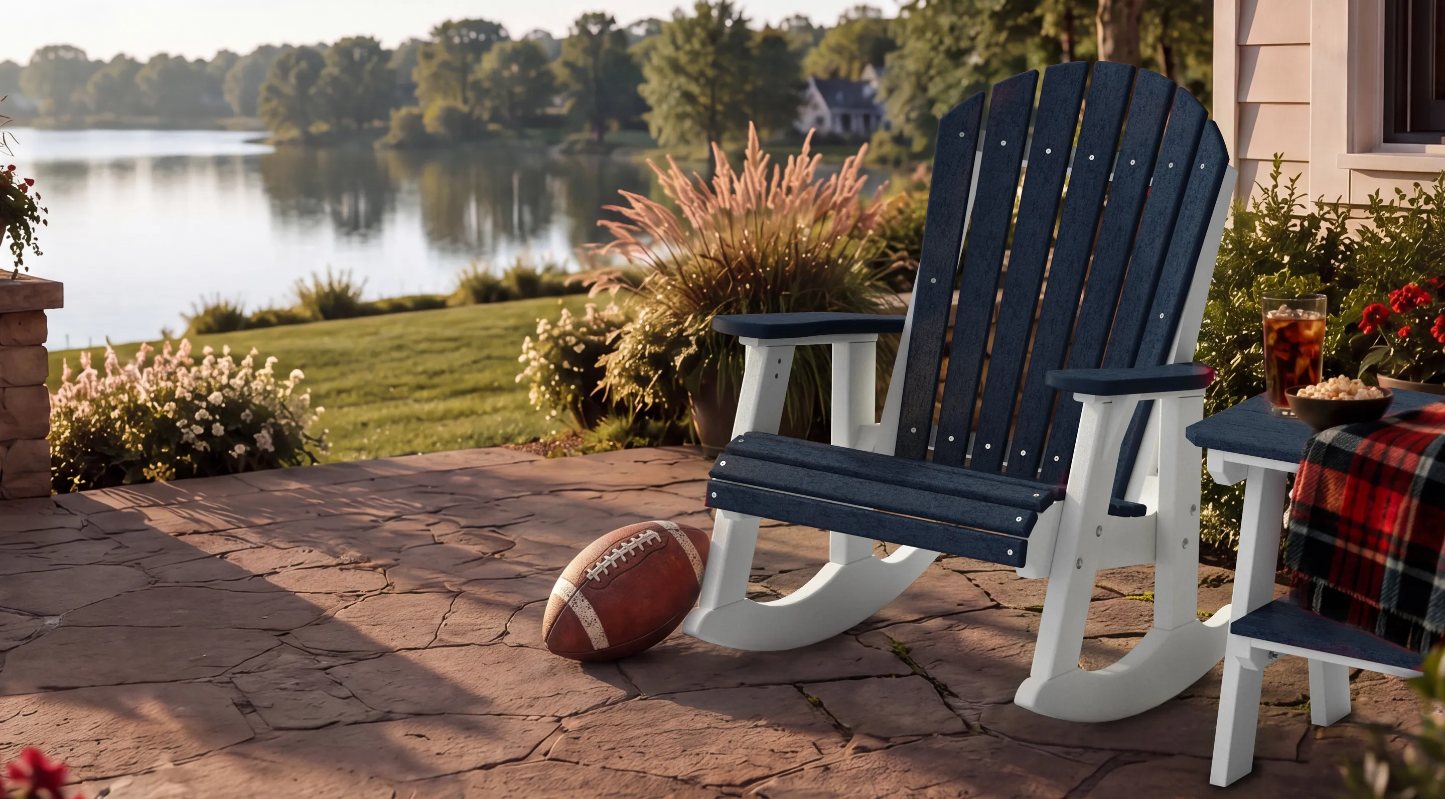 A navy and white Wildridge poly rocking chair on a flagstone patio overlooking a lake at golden hour, with a football, a plaid blanket and a side table alongside